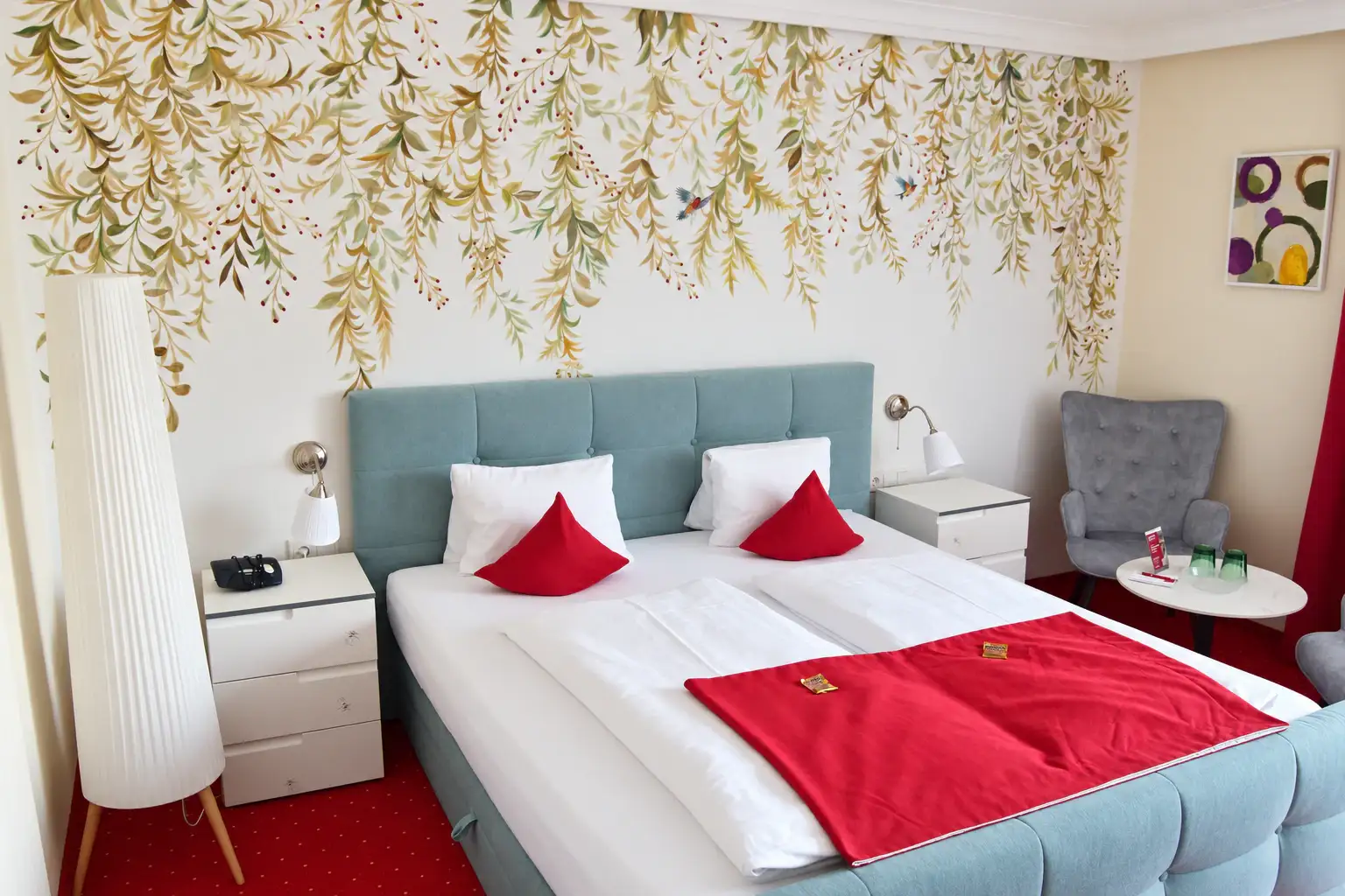 Doppelzimmer mit botanischer Wandgestaltung, gepolstertem Bett und roten Kissen