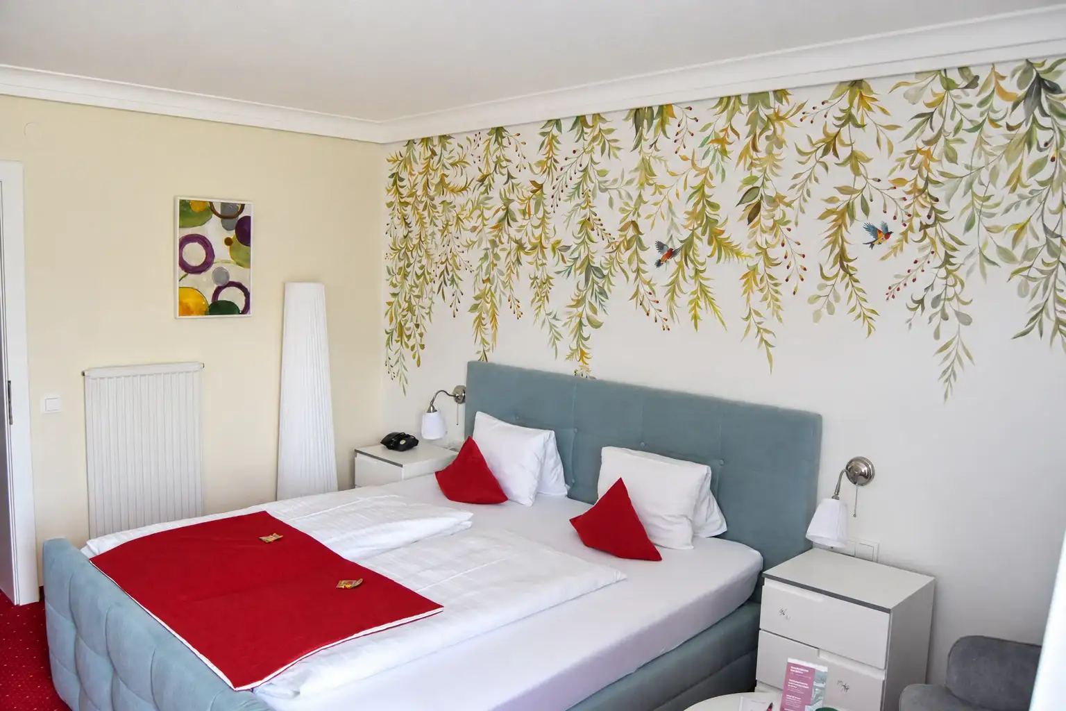 Zweite Ansicht eines Doppelzimmers mit gepolstertem Bett, botanischer Wandgestaltung und weißer Stehlampe
